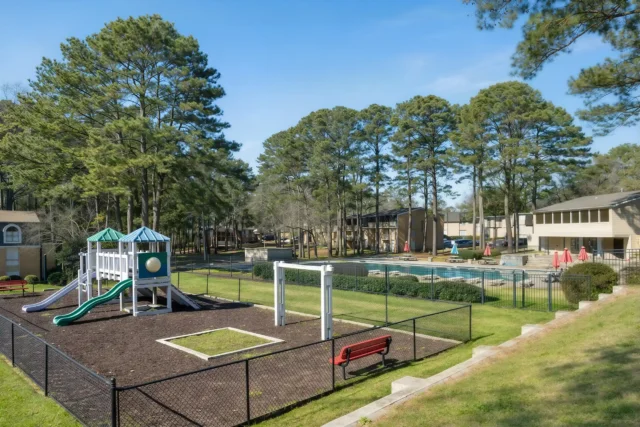 Fenced dog park at The Village on Riverwalk in College Park, GA, surrounded by mature trees with a glimpse of the apartment buildings in the background.