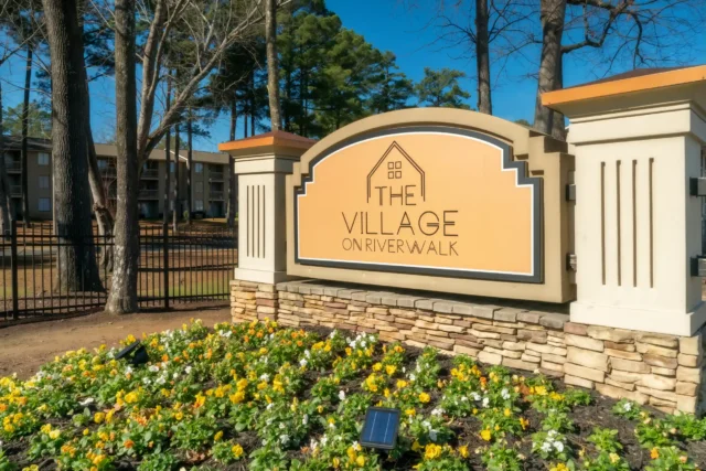 Monument sign for The Village on Riverwalk apartments in College Park, GA, surrounded by professional landscaping, mature trees, and residential buildings.